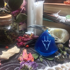 May include: A blue glass pendant with a white stylized design of a three-pronged symbol. The pendant is surrounded by various objects, including a bird skull, dried flowers, and a glass jar filled with moss.