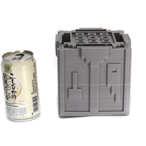 May include: A gray 3D printed cube with a series of holes on the top and a design on the front. The cube is next to a can of beer.