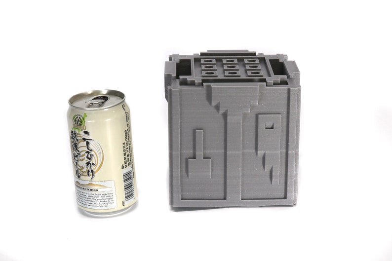 May include: A gray 3D printed cube with a series of holes on the top and a design on the front. The cube is next to a can of beer.