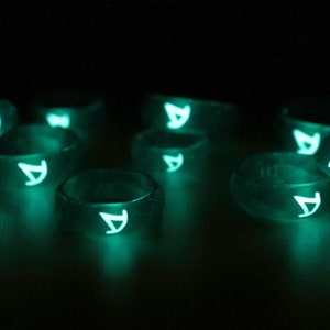 Pu&ograve; includere: Un gruppo di anelli che brillano al buio con una lettera "A" bianca su ogni anello. Gli anelli sono fatti di un materiale trasparente e traslucido e brillano di verde al buio.