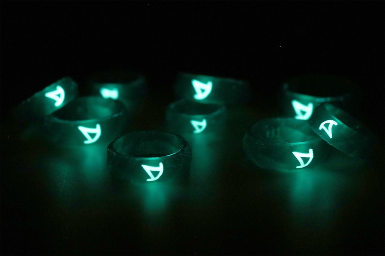 Pu&ograve; includere: Un gruppo di anelli che brillano al buio con una lettera "A" bianca su ogni anello. Gli anelli sono fatti di un materiale trasparente e traslucido e brillano di verde al buio.