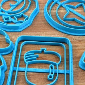 May include: A set of blue plastic cookie cutters in various shapes, including a star, an acorn, a snowman, and a swirl design. The cutters are arranged on a wooden surface.