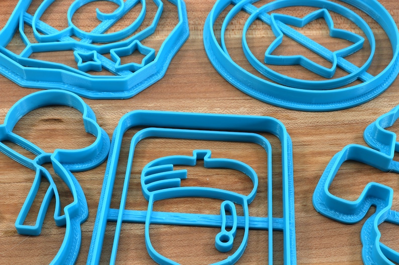 May include: A set of blue plastic cookie cutters in various shapes, including a star, an acorn, a snowman, and a swirl design. The cutters are arranged on a wooden surface.