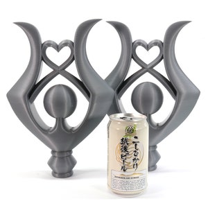 May include: Two silver-colored metal sculptures with a heart design and a sphere in the center. The sculptures are standing on a white surface next to a can of Japanese green tea.