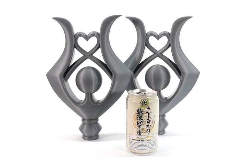 May include: Two silver-colored metal sculptures with a heart design and a sphere in the center. The sculptures are standing on a white surface next to a can of Japanese green tea.