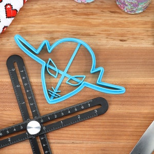 May include: A blue plastic cookie cutter shaped like a cartoon alien head with a single eye and two antennas. The cutter is on a wooden surface with a black measuring tool.