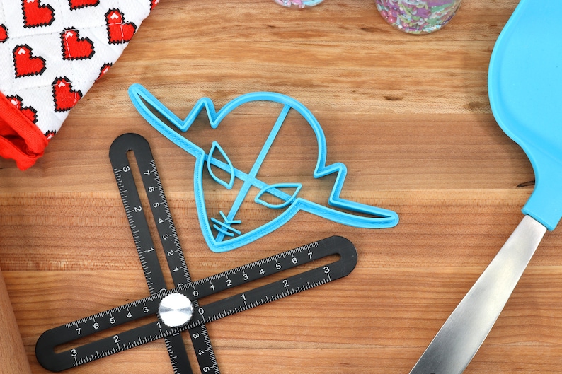 May include: A blue plastic cookie cutter shaped like a cartoon alien head with a single eye and two antennas. The cutter is on a wooden surface with a black measuring tool.