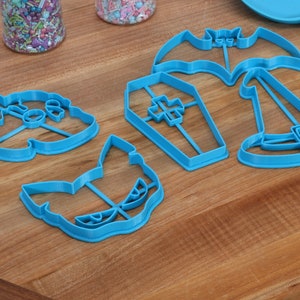 May include: A set of five blue plastic Halloween cookie cutters. The cutters are shaped like a bat, a coffin, a witch's hat, a ghost, and a cat.