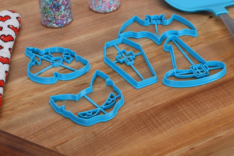 May include: A set of five blue plastic Halloween cookie cutters. The cutters are shaped like a bat, a coffin, a witch's hat, a ghost, and a cat.