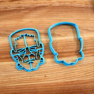 May include: Two blue plastic cookie cutters, one shaped like a Frankenstein's monster face and the other shaped like a simple oval.