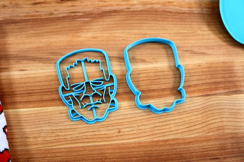 May include: Two blue plastic cookie cutters, one shaped like a Frankenstein's monster face and the other shaped like a simple oval.