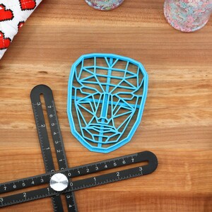 May include: A blue plastic face-shaped cookie cutter with a geometric design. The cutter is on a wooden surface with a black ruler and a white and red fabric.