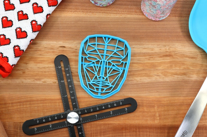 May include: A blue plastic face-shaped cookie cutter with a geometric design. The cutter is on a wooden surface with a black ruler and a white and red fabric.