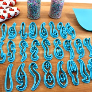 May include: A set of 26 blue plastic cookie cutters shaped like the letters of the alphabet in a stylized font. The cutters are arranged in three rows on a wooden surface.