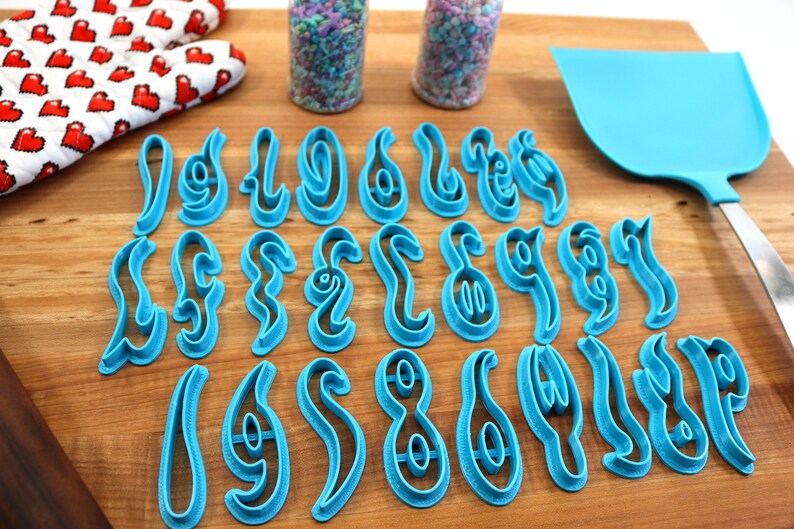 May include: A set of 26 blue plastic cookie cutters shaped like the letters of the alphabet in a stylized font. The cutters are arranged in three rows on a wooden surface.