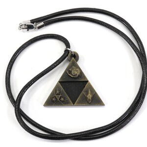 May include: A black cord necklace with a gold-toned pendant featuring a triangular design with symbols. The pendant is a stylized representation of the Triforce from the Legend of Zelda video game series.