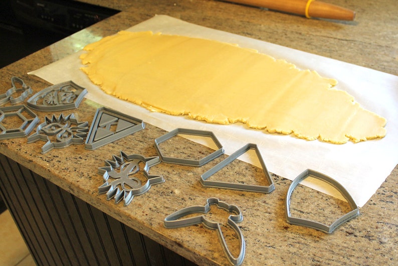 Puede incluir: Una hoja de masa para galletas sobre papel de hornear con varios cortadores de galletas en una encimera. Los cortadores de galletas tienen forma de hex&aacute;gono, tri&aacute;ngulo, mariposa, escudo y corona.