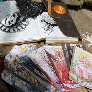 May include: A leather-bound journal with a black and white illustration of a celestial map and a set of tarot cards spread out on a wooden surface. The cards feature colorful illustrations of angels and mythical creatures.