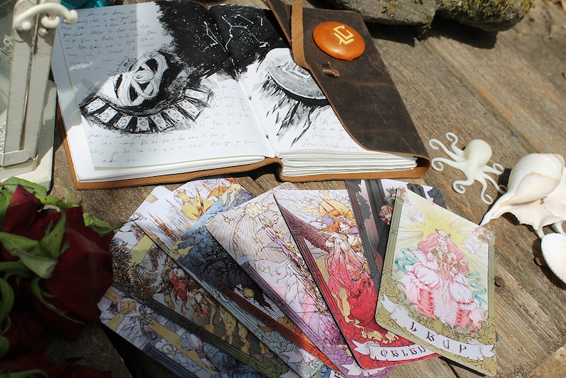 May include: A leather-bound journal with a black and white illustration of a celestial map and a set of tarot cards spread out on a wooden surface. The cards feature colorful illustrations of angels and mythical creatures.
