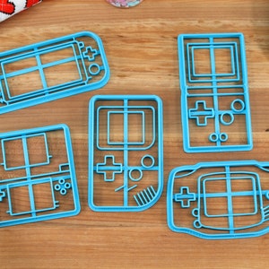 May include: Five blue plastic cookie cutters shaped like different handheld video game consoles.