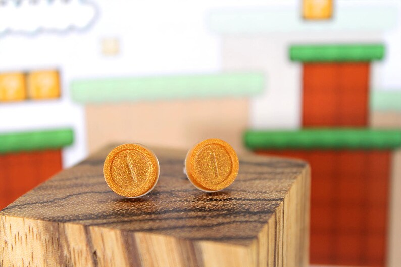 May include: A pair of gold-colored earrings shaped like a video game coin. The earrings are on a wooden surface.