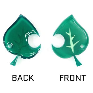 May include: A green leaf-shaped charm with a white leaf design on the front and a smooth surface on the back.
