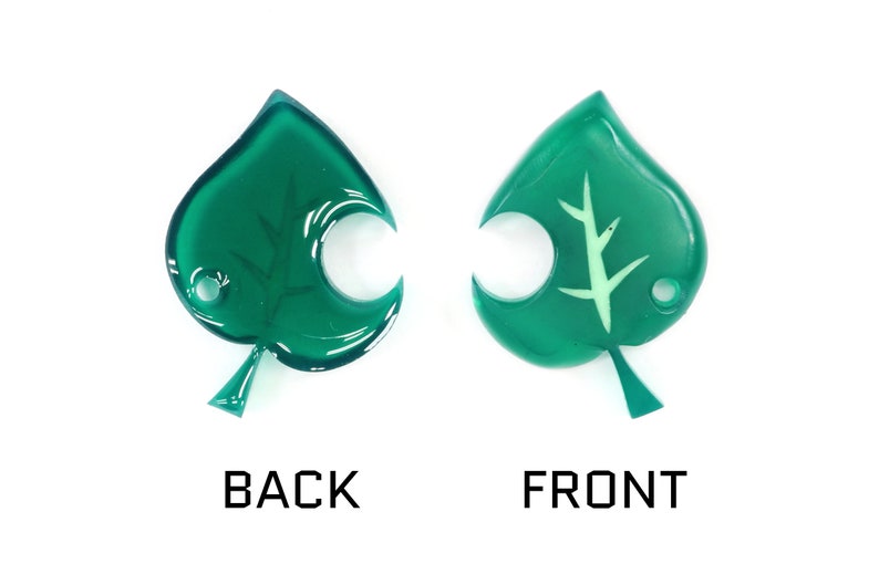 May include: A green leaf-shaped charm with a white leaf design on the front and a smooth surface on the back.