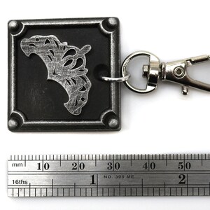 May include: A silver metal keychain with a black square charm featuring a silver filigree design. The charm is attached to a silver metal swivel clasp.