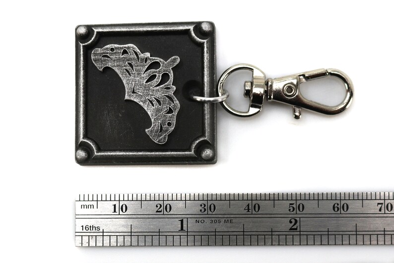 May include: A silver metal keychain with a black square charm featuring a silver filigree design. The charm is attached to a silver metal swivel clasp.