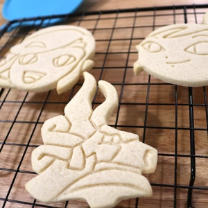 May include: Three sugar cookies on a black wire cooling rack. The cookies are in the shape of a cartoon face, a dragon, and a cartoon face with a ponytail.