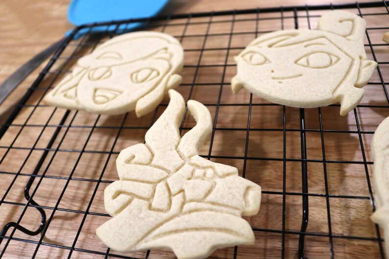May include: Three sugar cookies on a black wire cooling rack. The cookies are in the shape of a cartoon face, a dragon, and a cartoon face with a ponytail.