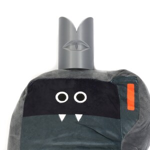 May include: Grey plush toy with a black face, two white eyes, and a white mouth with two fangs. The toy has a grey felt crown with a white eye on the top.