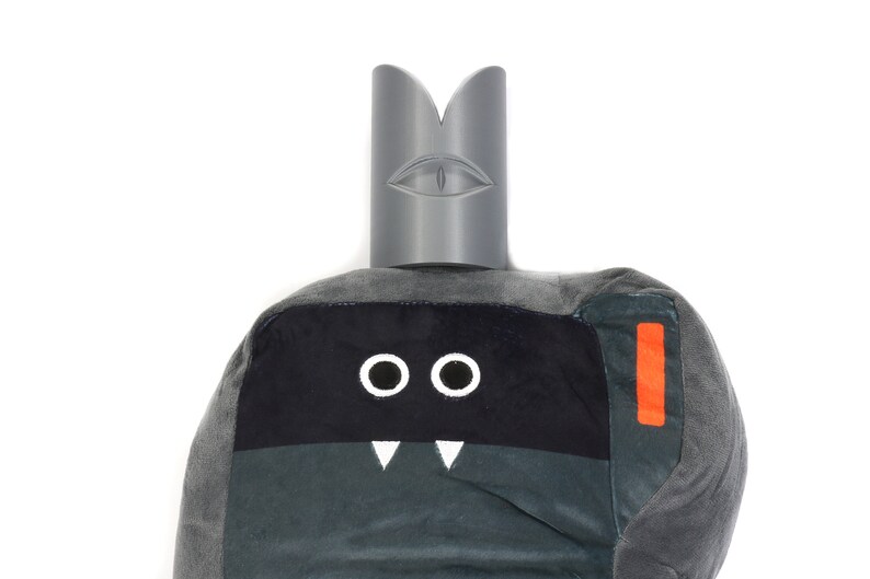 May include: Grey plush toy with a black face, two white eyes, and a white mouth with two fangs. The toy has a grey felt crown with a white eye on the top.