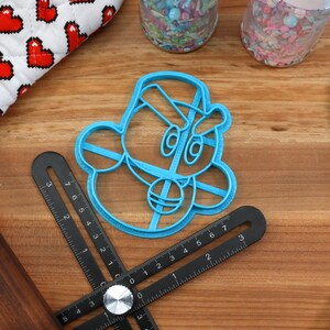 May include: A blue plastic cookie cutter shaped like a cartoon character with a hat and a large nose. The cutter is on a wooden surface with a ruler and a rolling pin.