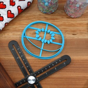 May include: A blue plastic cookie cutter shaped like a circle with a design of a sun with rays. The cutter is on a wooden surface with a black ruler and a rolling pin.