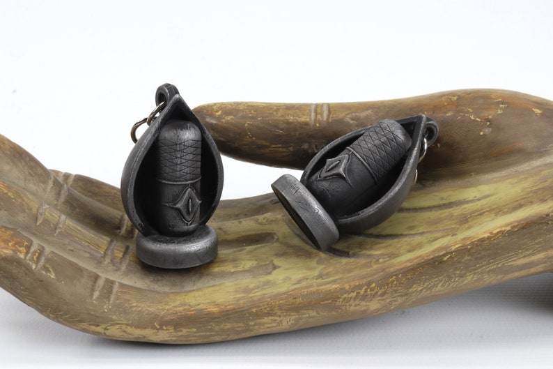 May include: Two silver-colored pendant earrings shaped like a teardrop with a small, detailed, black, cylindrical charm inside. The charms have a geometric design.