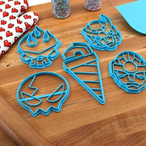 May include: A set of six blue plastic cookie cutters shaped like a skull with flames, a drill, a skull with a lightning bolt, and two different faces. The cutters are on a wooden surface.