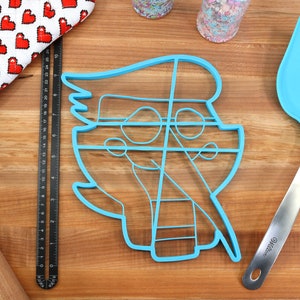 May include: A blue plastic cookie cutter shaped like a cartoon character with a large head and a small body. The cutter has a detailed design with lines and circles. The cutter is on a wooden surface with a rolling pin, a spatula, and a measuring tape.