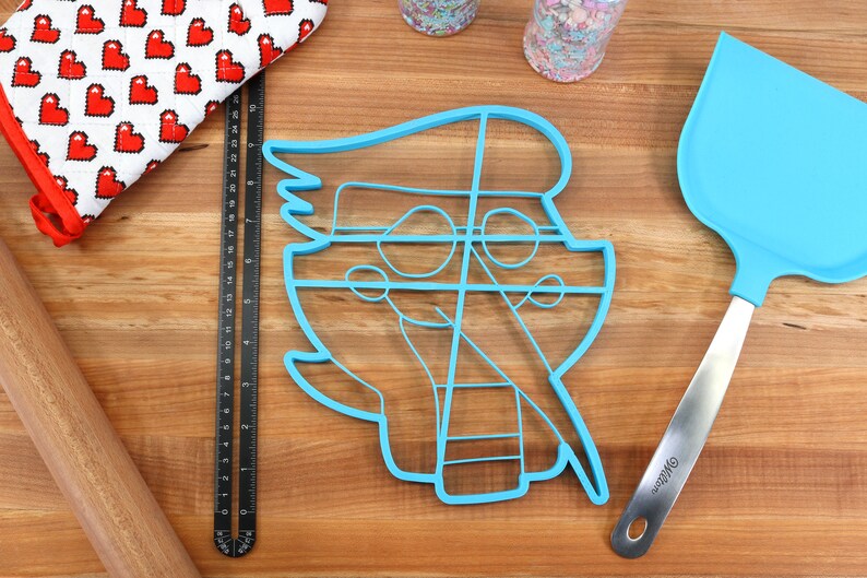 May include: A blue plastic cookie cutter shaped like a cartoon character with a large head and a small body. The cutter has a detailed design with lines and circles. The cutter is on a wooden surface with a rolling pin, a spatula, and a measuring tape.