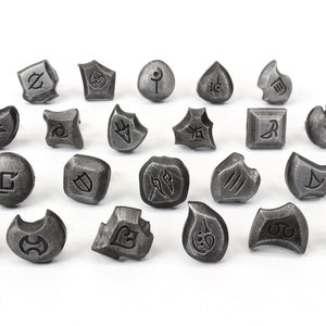 Puede incluir: Una colecci&oacute;n de veinticuatro objetos grises, similares a piedras, con s&iacute;mbolos grabados. Los objetos var&iacute;an en forma, incluyendo formas geom&eacute;tricas y org&aacute;nicas. Cada pieza presenta un dise&ntilde;o &uacute;nico grabado, sugiriendo un conjunto de piezas de juego o elementos decorativos.
