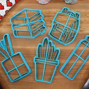 K&ouml;nnte beinhalten: F&uuml;nf blaue Plastik-Ausstecher in Form eines B&uuml;cherstapels, eines Rucksacks, einer Topfpflanze, einer Wasserflasche und einer Geschenkbox. Die Ausstecher liegen auf einer Holzoberfl&auml;che.