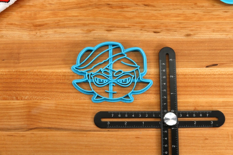 May include: A blue plastic cookie cutter shaped like a cartoon character with a black eye mask. The cookie cutter is on a wooden surface with a black ruler.