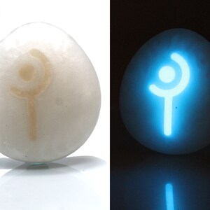 May include: A white egg-shaped stone with a glowing blue symbol of a staff with a circle on top. The symbol is visible in the dark.