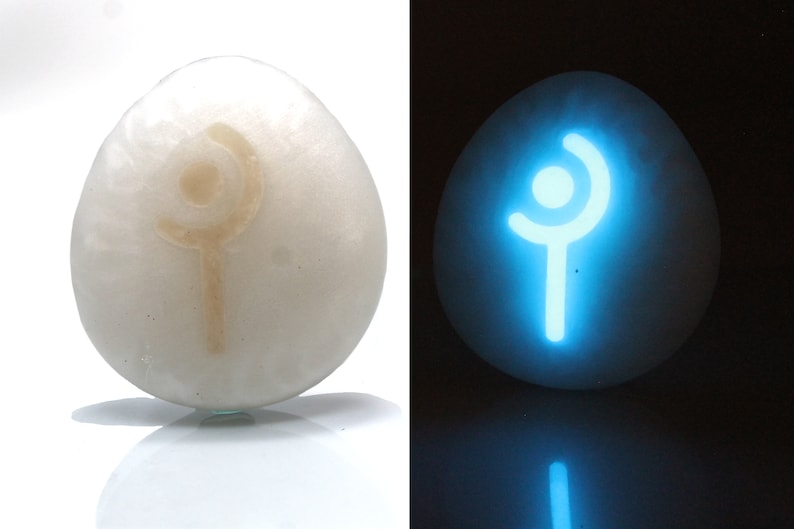 May include: A white egg-shaped stone with a glowing blue symbol of a staff with a circle on top. The symbol is visible in the dark.