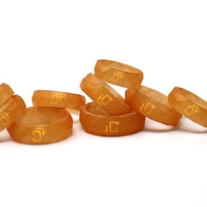 May include: A group of eight orange resin rings with a geometric design and a gold inscription. The rings are stacked on top of each other.