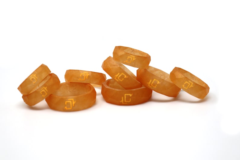 May include: A group of eight orange resin rings with a geometric design and a gold inscription. The rings are stacked on top of each other.