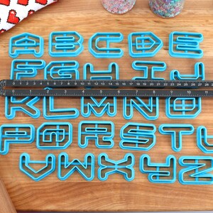 May include: A set of 26 blue plastic alphabet cookie cutters on a wooden surface. The cutters are arranged in alphabetical order and are approximately 2 inches tall.