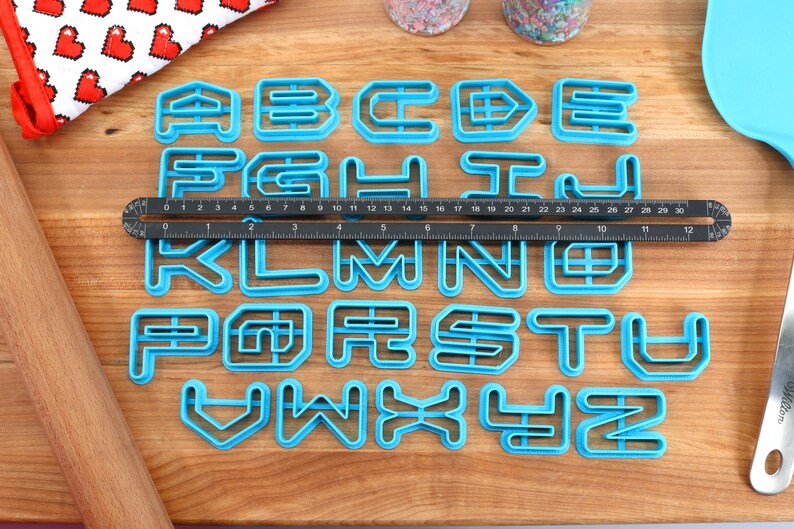 May include: A set of 26 blue plastic alphabet cookie cutters on a wooden surface. The cutters are arranged in alphabetical order and are approximately 2 inches tall.