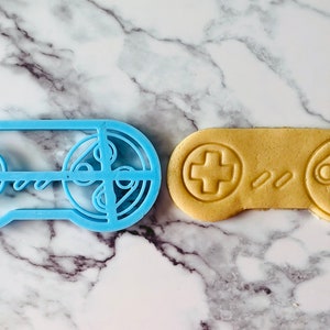 May include: A blue plastic cookie cutter shaped like a video game controller next to a light brown cookie in the shape of a video game controller.