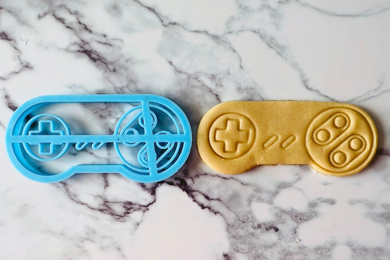 May include: A blue plastic cookie cutter shaped like a video game controller next to a light brown cookie in the shape of a video game controller.